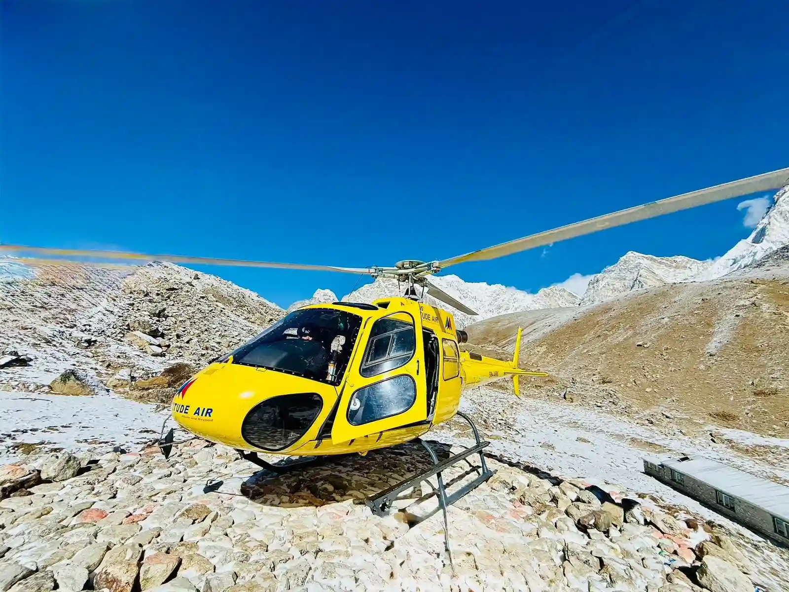 An eye-level medium shot captures a bright yellow Altitude Air helicopter landed on a rugged, rocky plateau in the Himalayas.